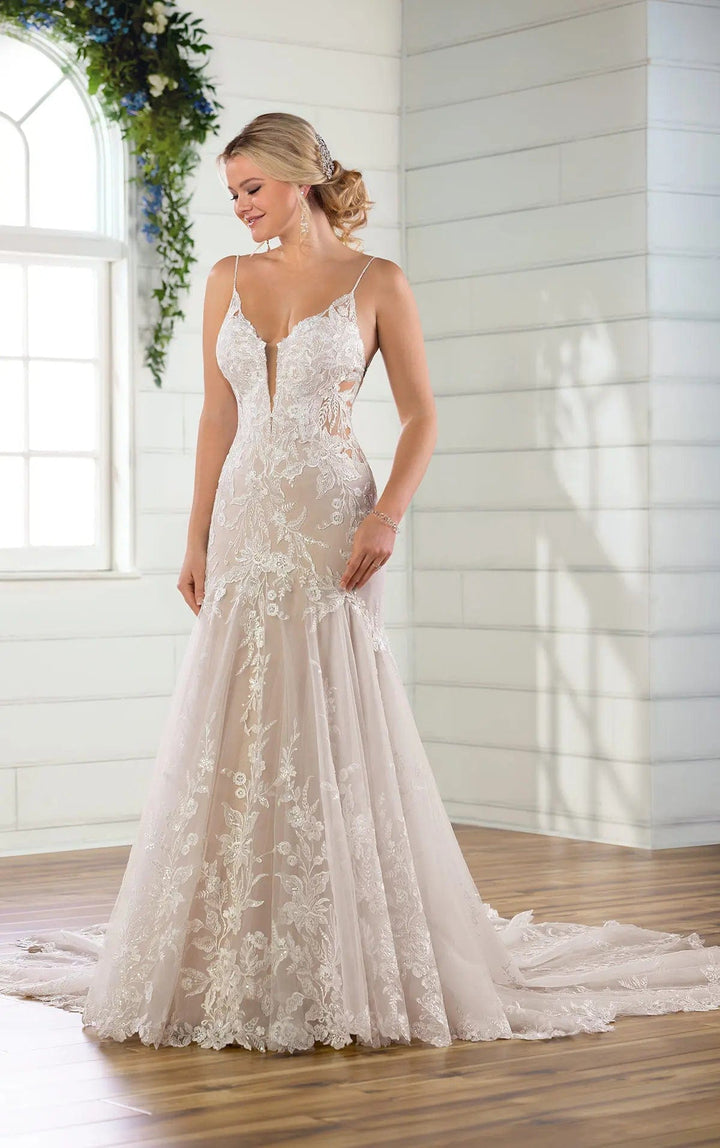 A woman modeling a sleeveless ivory wedding gown with floral lace applique, a deep V-neckline, and a full mermaid-style skirt with a long train in a bright indoor room with white paneled walls and wood flooring.