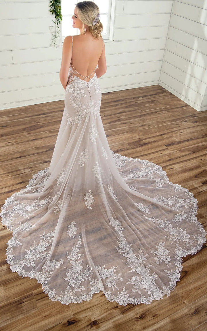 A woman stands with her back to the camera wearing a sleeveless lace wedding gown with a long train in a bright room with white paneled walls and a wood floor.