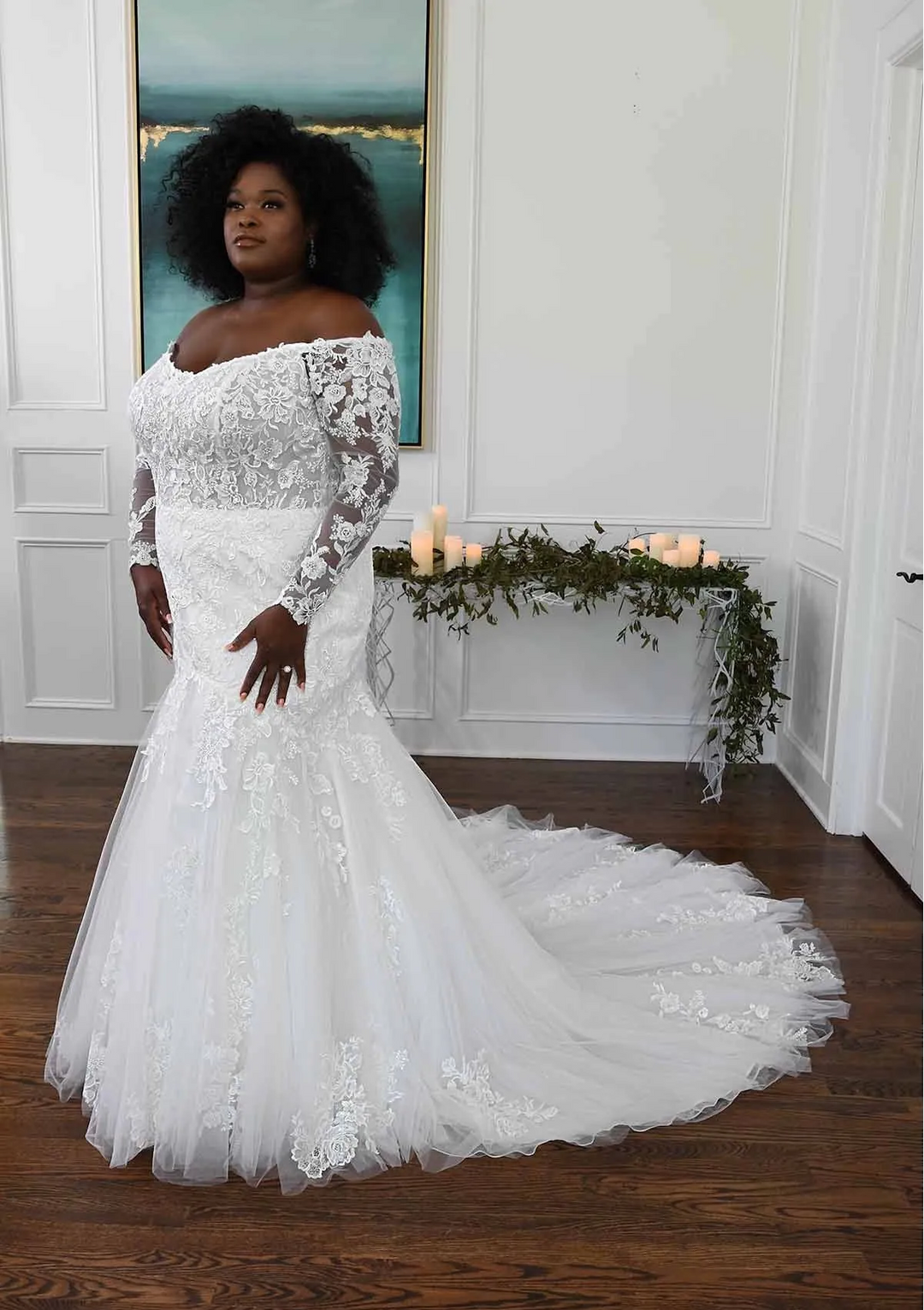 Woman in a white lace wedding dress standing in a room with candles and greenery.