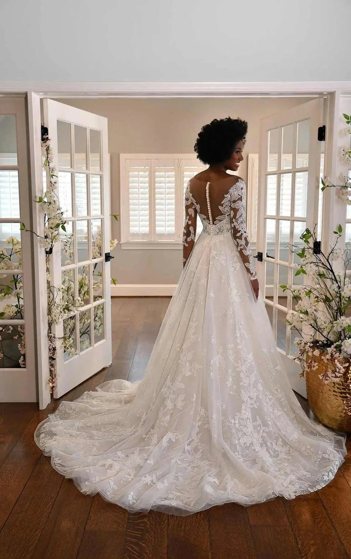 A woman wearing a white lace wedding gown with long sleeves and an open back, standing in a doorway with a long train spread on a wooden floor.