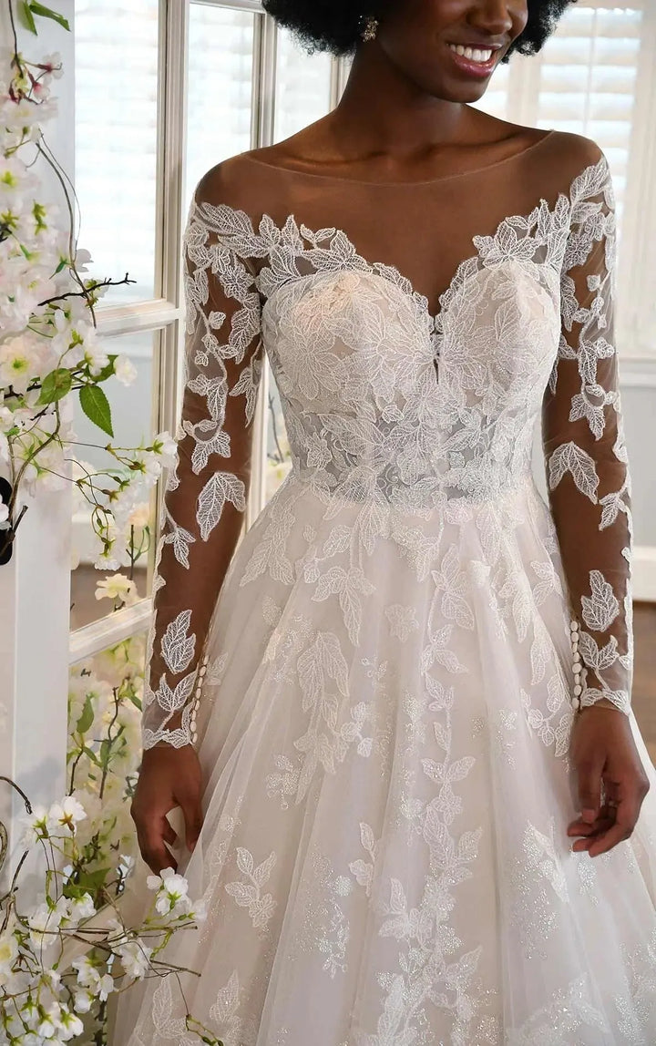 A woman wearing a white lace wedding dress with long sheer sleeves, a sweetheart neckline, and a full tulle skirt, photographed indoors near a window and floral decor.
