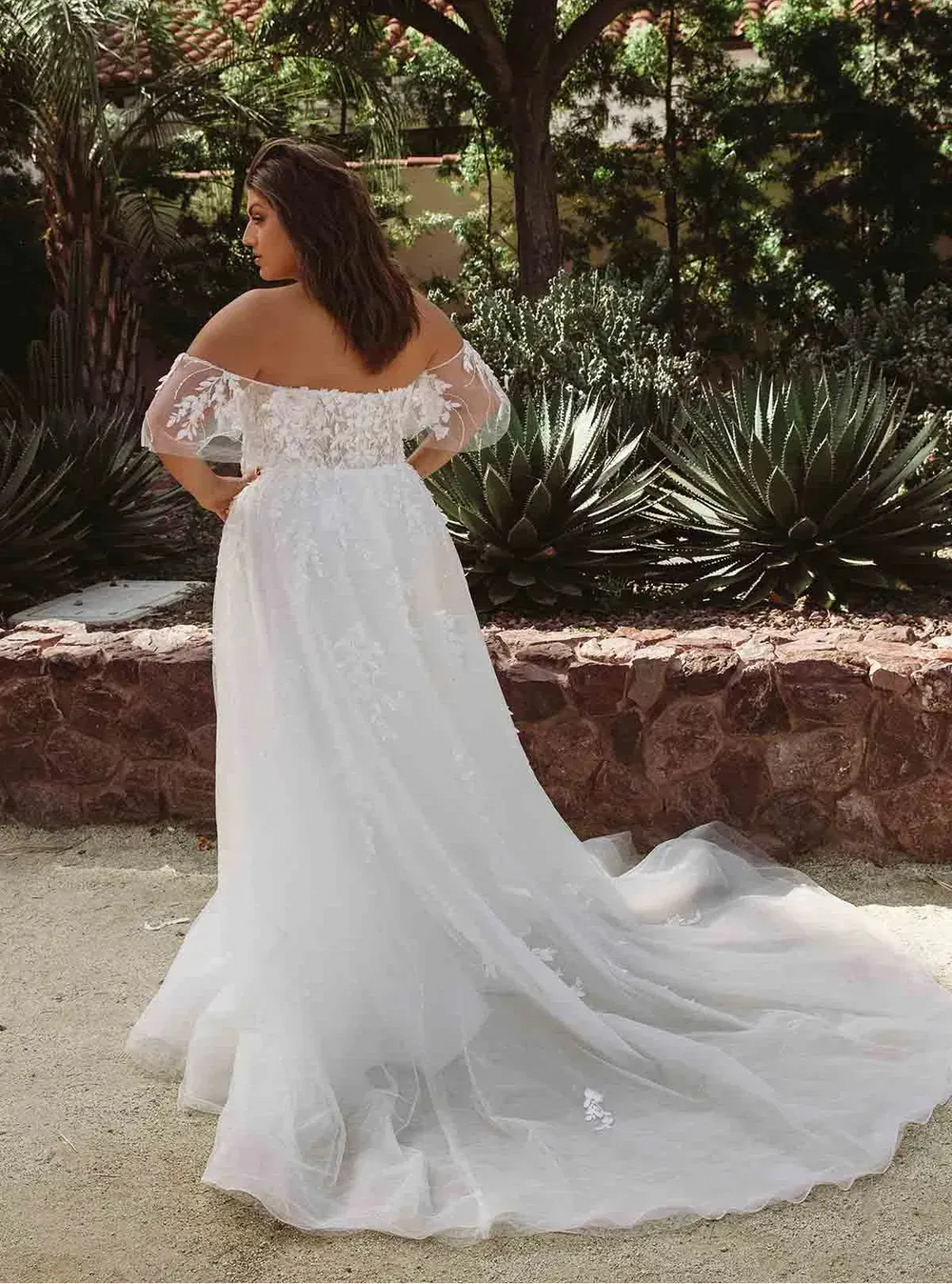 Woman in a white off-shoulder wedding dress standing outdoors with plants in the background