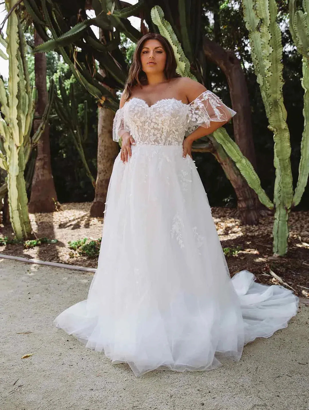 A woman wearing a white off-the-shoulder A-line wedding gown with lace applique and a tulle skirt, standing outdoors in front of cacti and trees.