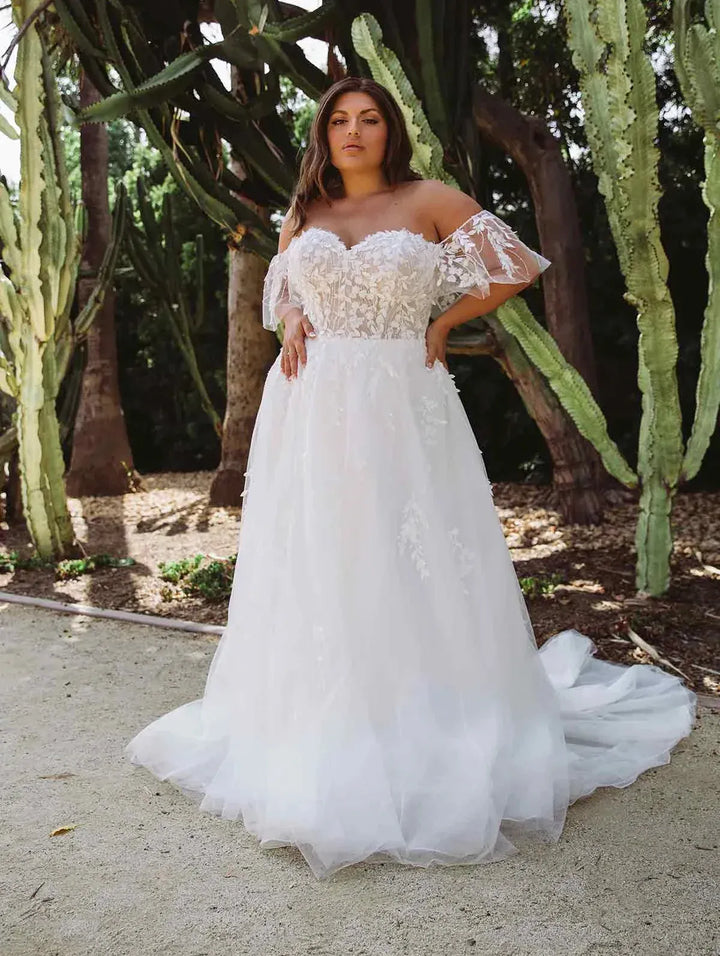 A woman wearing a white off-the-shoulder A-line wedding gown with lace applique and a tulle skirt, standing outdoors in front of cacti and trees.