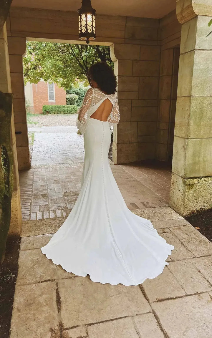 A woman wearing a white mermaid-style wedding dress with a lace-trimmed back and long sleeves, standing in a stone doorway and facing away from the camera.