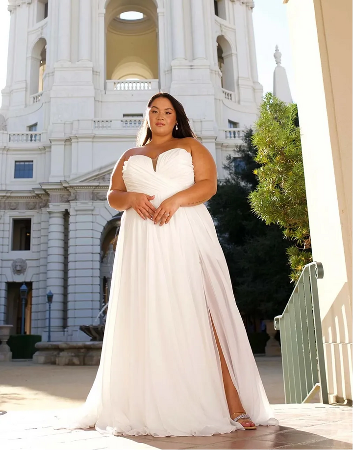 A woman wearing a strapless white wedding gown with a sweetheart neckline and a high side slit, standing in front of a large white domed building framed by an archway.