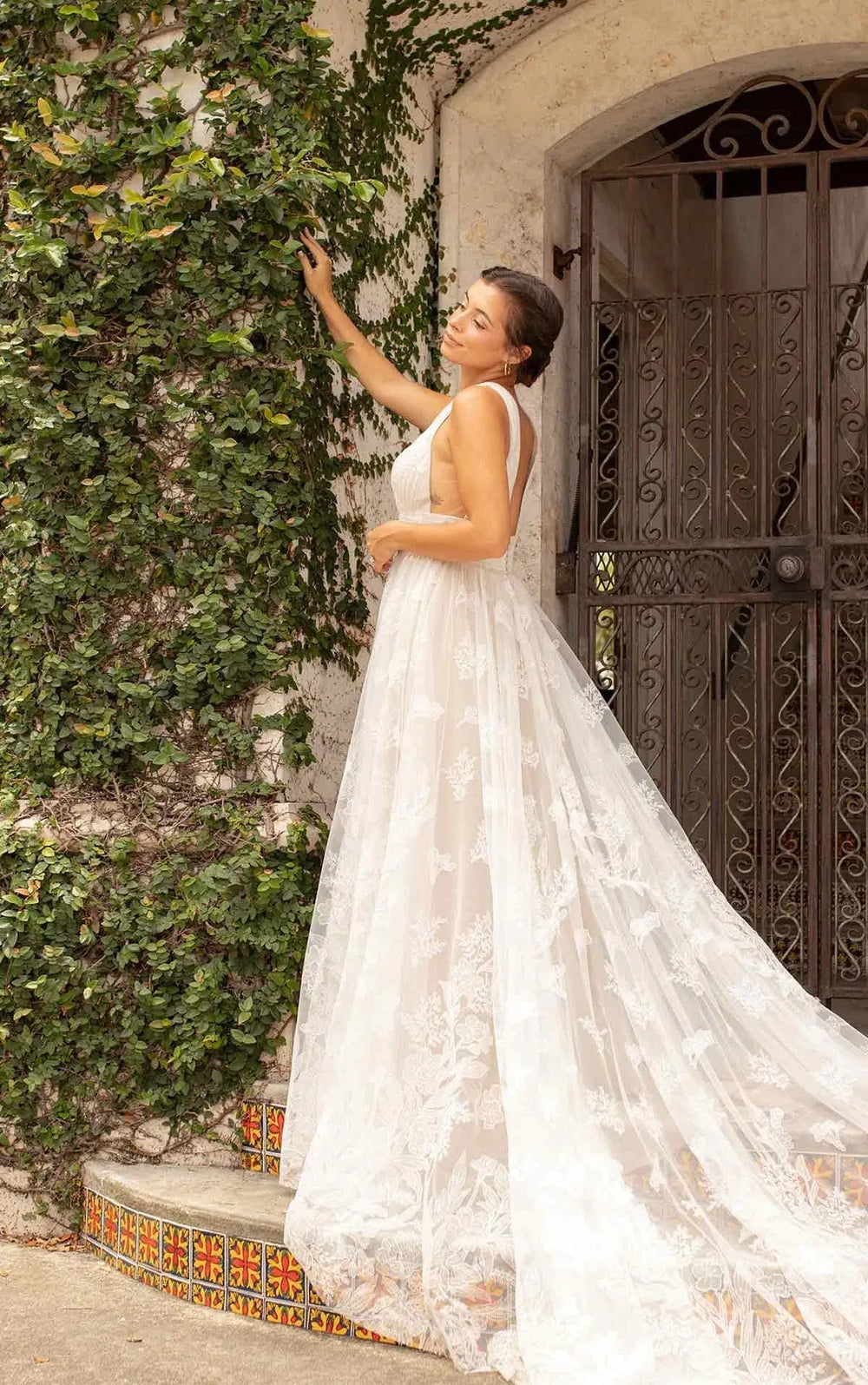 Woman in a white lace dress standing next to a textured wall with greenery.