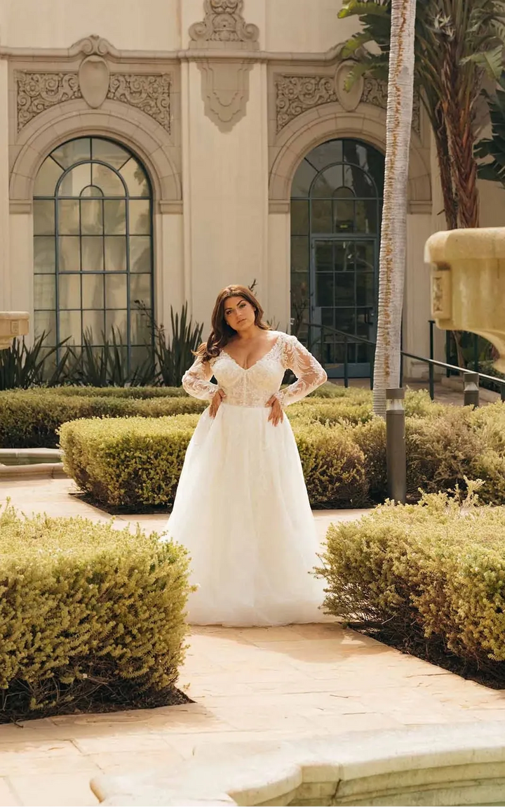 A woman wearing a white lace wedding gown stands in a landscaped courtyard with trimmed hedges, palm trees, and a beige stucco building in the background.