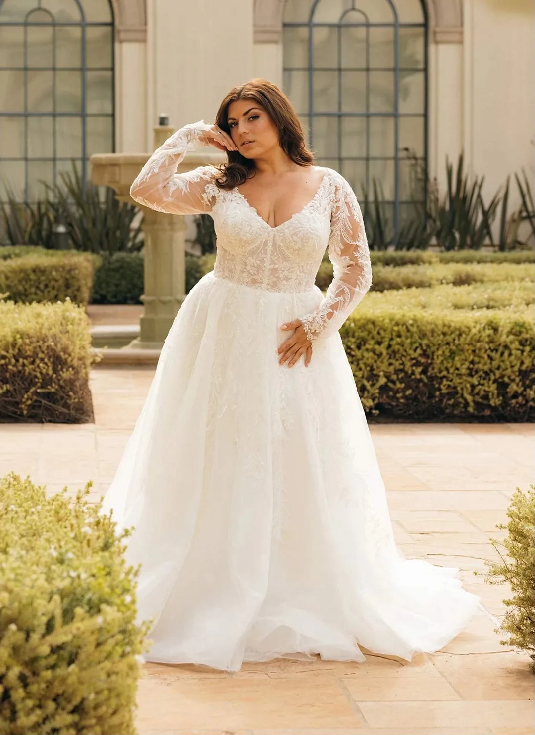 A woman wearing a white lace wedding gown with long sleeves and a full skirt, standing in a garden courtyard with trimmed hedges and large arched windows in the background.
