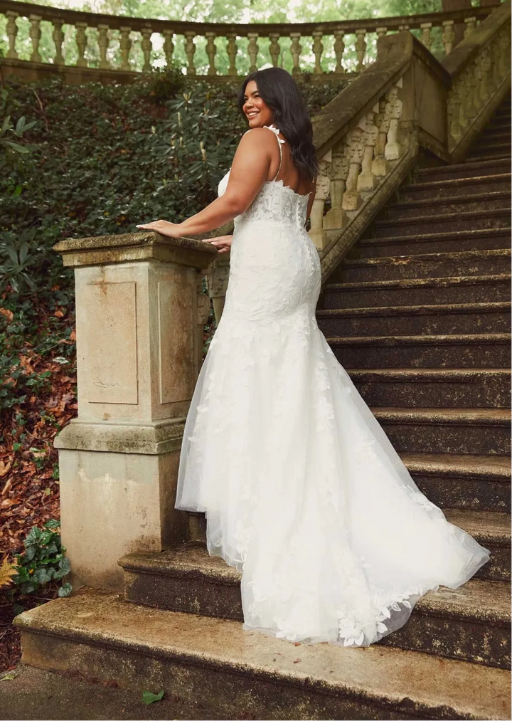 A woman wearing a white lace mermaid-style wedding gown stands on outdoor stone stairs beside a balustrade, photographed in a garden setting.