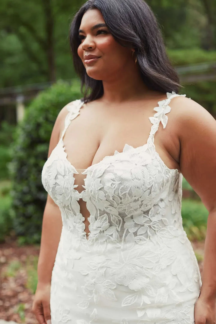 A woman wearing a white floral lace wedding dress with thin straps and a deep V-neckline, photographed outdoors against a blurred green garden background.