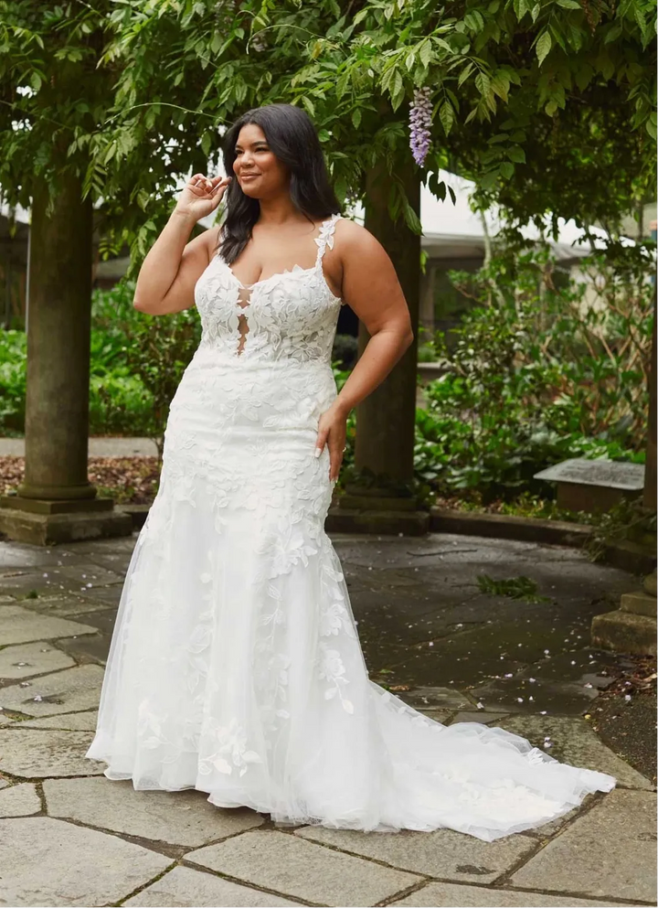 A woman wearing a white lace wedding gown stands outdoors on a stone patio beneath leafy trees, with the dress showing a fitted bodice, lace appliqué, and a long train.