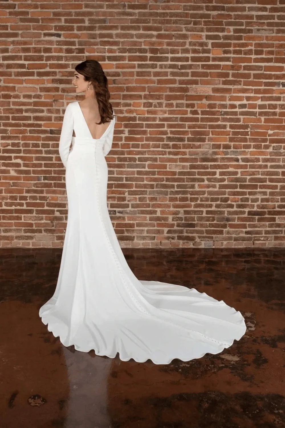 A woman wearing a white wedding gown stands in profile against a red-brown brick wall, with the long train of the dress pooling on the floor.