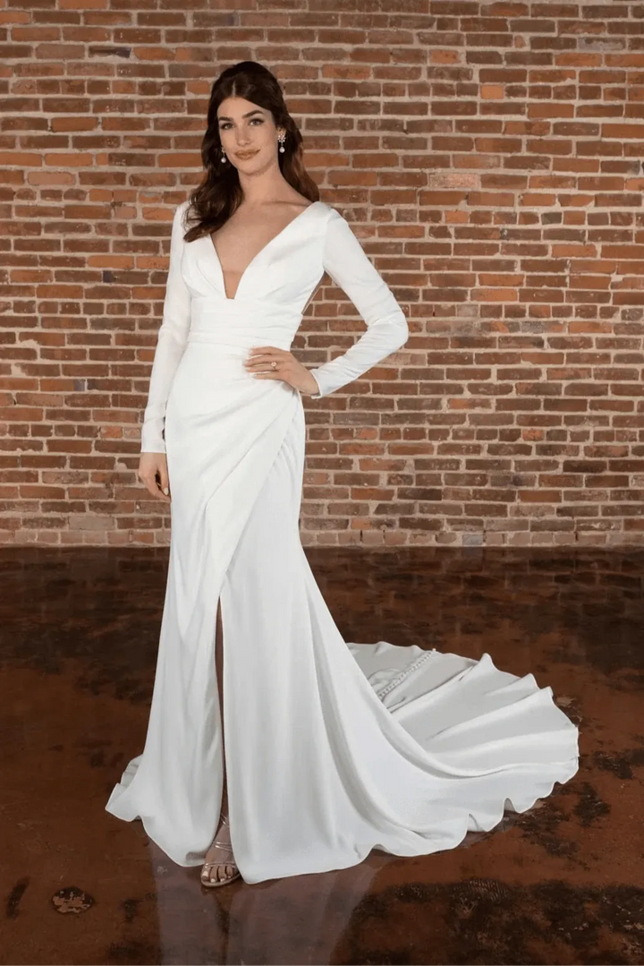 A woman modeling a long-sleeve white wedding gown with a deep V-neckline and a slight train, standing in front of a red-brown brick wall.