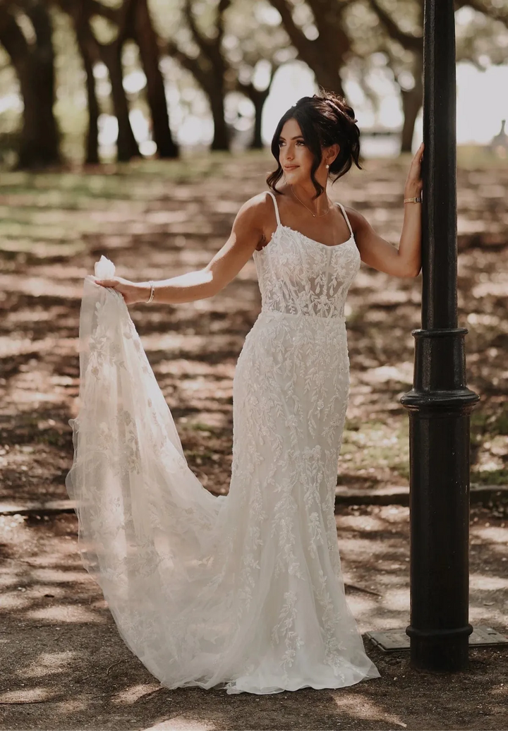 A woman wearing a sleeveless lace wedding gown stands outdoors beside a black pole, holding out the train of the dress in a park-like setting with trees and dappled sunlight.