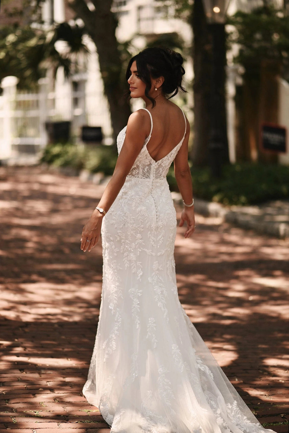 A woman wearing a white lace wedding gown with a low open back and spaghetti straps, photographed outdoors on a sun-dappled brick path with trees and a building in the background.
