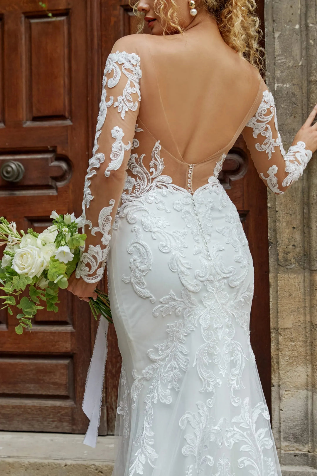 Woman wearing a white lace wedding dress with long sleeves, standing in front of a wooden door.
