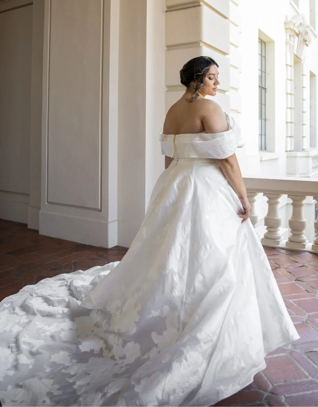 Woman in a white wedding dress standing in an elegant indoor setting with columns and light.