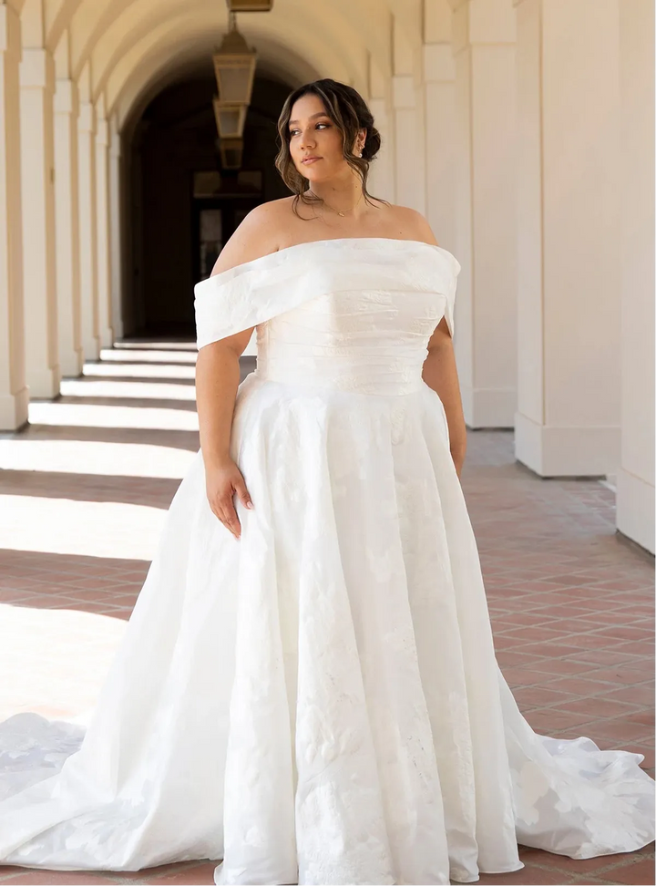 Woman in a white off-shoulder wedding dress standing in an architectural hallway.