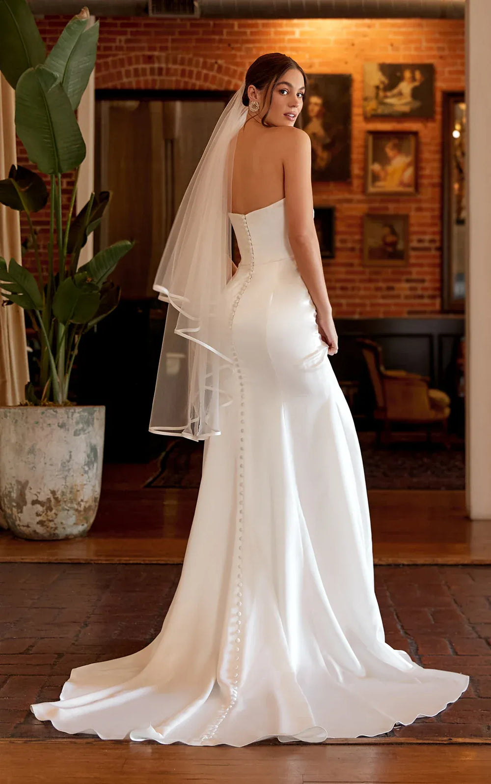 Woman in a white wedding dress with a veil standing in a room with brick walls and framed pictures.