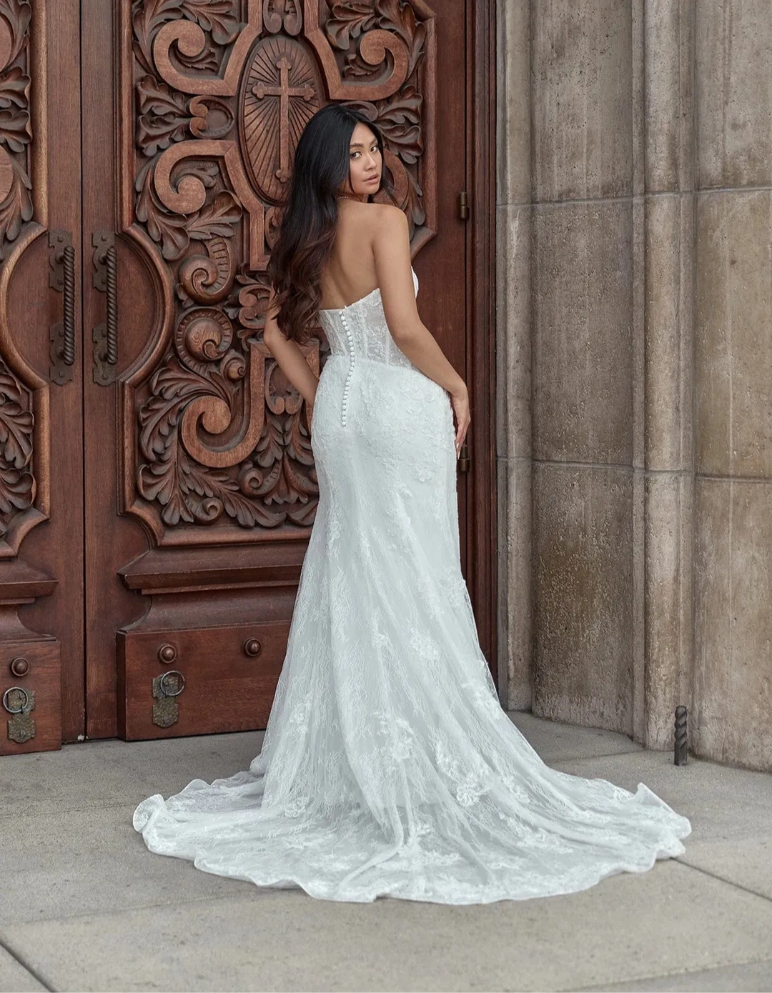 A woman wearing a strapless white lace wedding gown stands in front of ornate carved wooden doors and a stone wall, photographed outdoors.