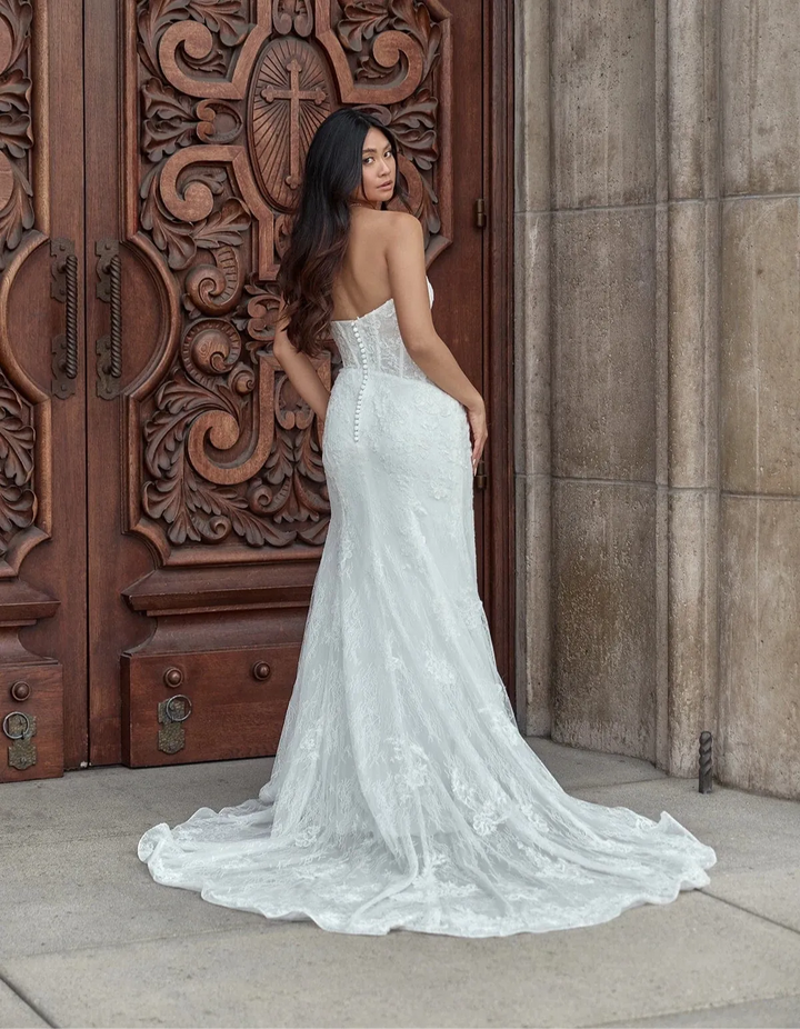 A woman wearing a strapless white lace wedding gown stands in front of ornate carved wooden doors and a stone wall, photographed outdoors.