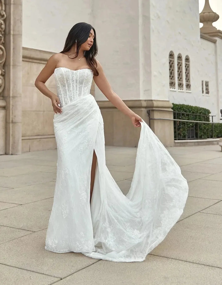 A woman wearing a strapless white lace wedding gown with a fitted bodice, draped skirt, and long train, standing outdoors in front of a beige stone building.