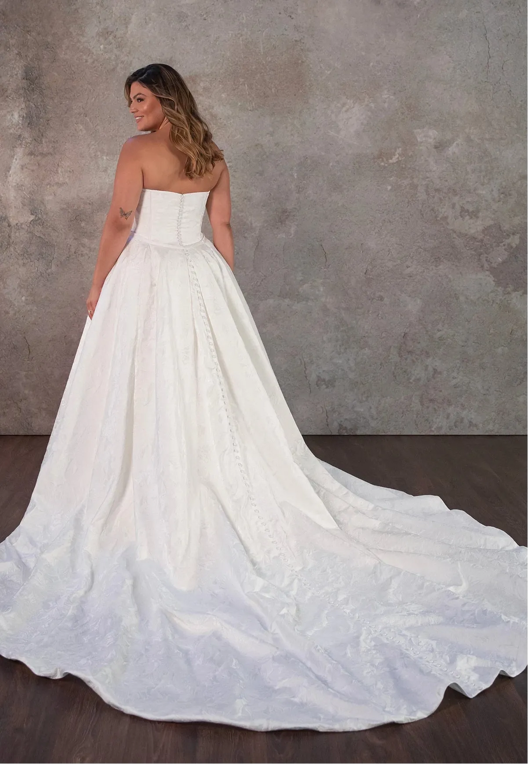 Woman wearing a strapless white wedding gown with a long train against a textured gray wall.