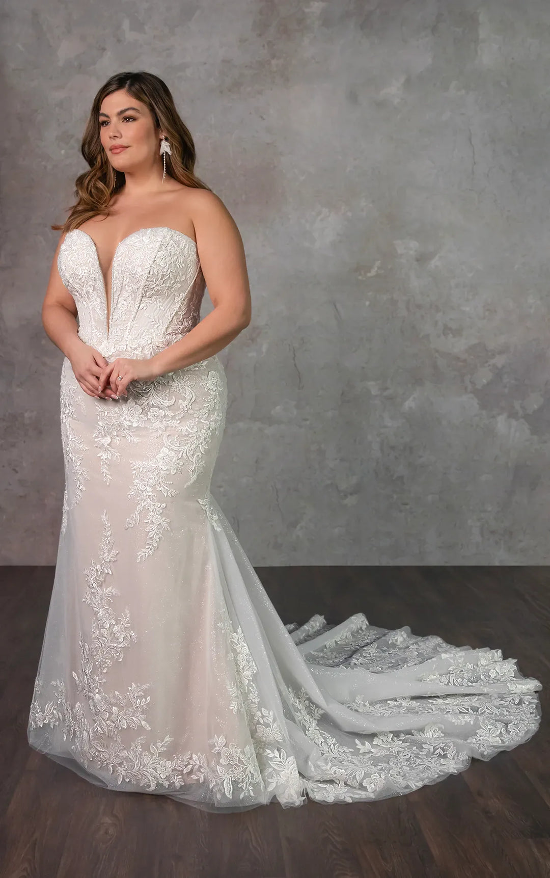 A woman modeling a strapless white lace wedding gown with a deep V neckline, fitted silhouette, and long train against a textured gray studio backdrop.