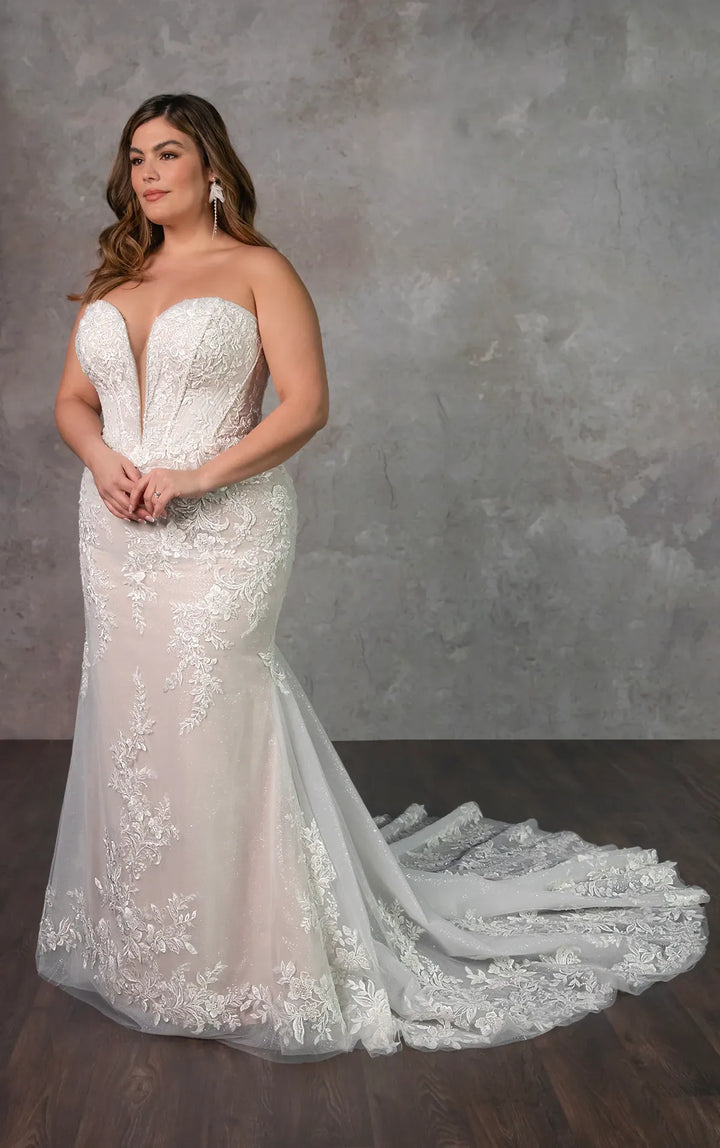 A woman modeling a strapless white lace wedding gown with a deep V neckline, fitted silhouette, and long train against a textured gray studio backdrop.