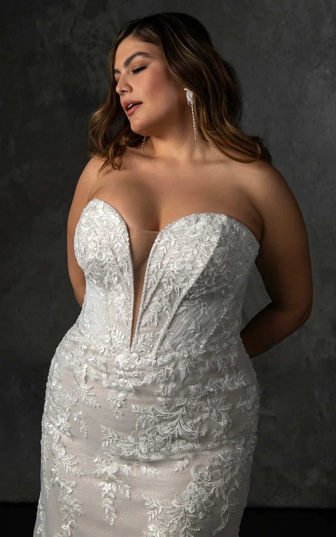 A woman modeling a strapless white lace wedding gown with a deep V-neckline, photographed against a dark textured studio background.