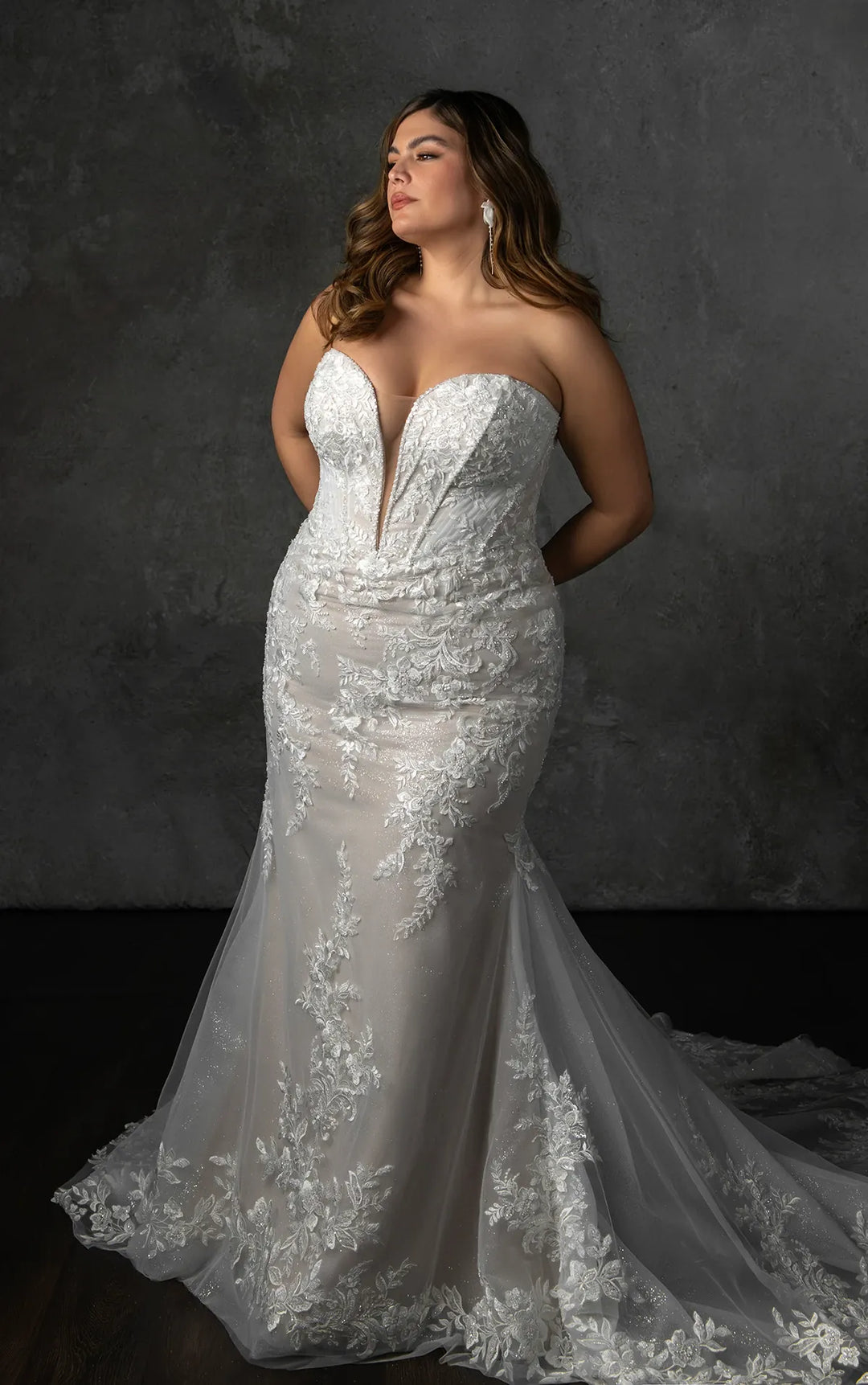 A woman modeling a strapless white lace mermaid-style wedding gown with a deep V neckline and a long train, posed against a dark textured studio backdrop.