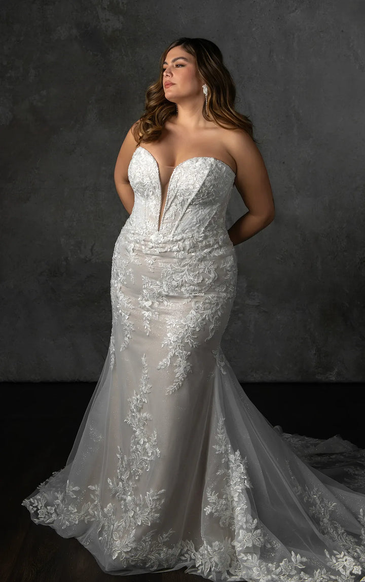 A woman modeling a strapless white lace mermaid-style wedding gown with a deep V neckline and a long train, posed against a dark textured studio backdrop.