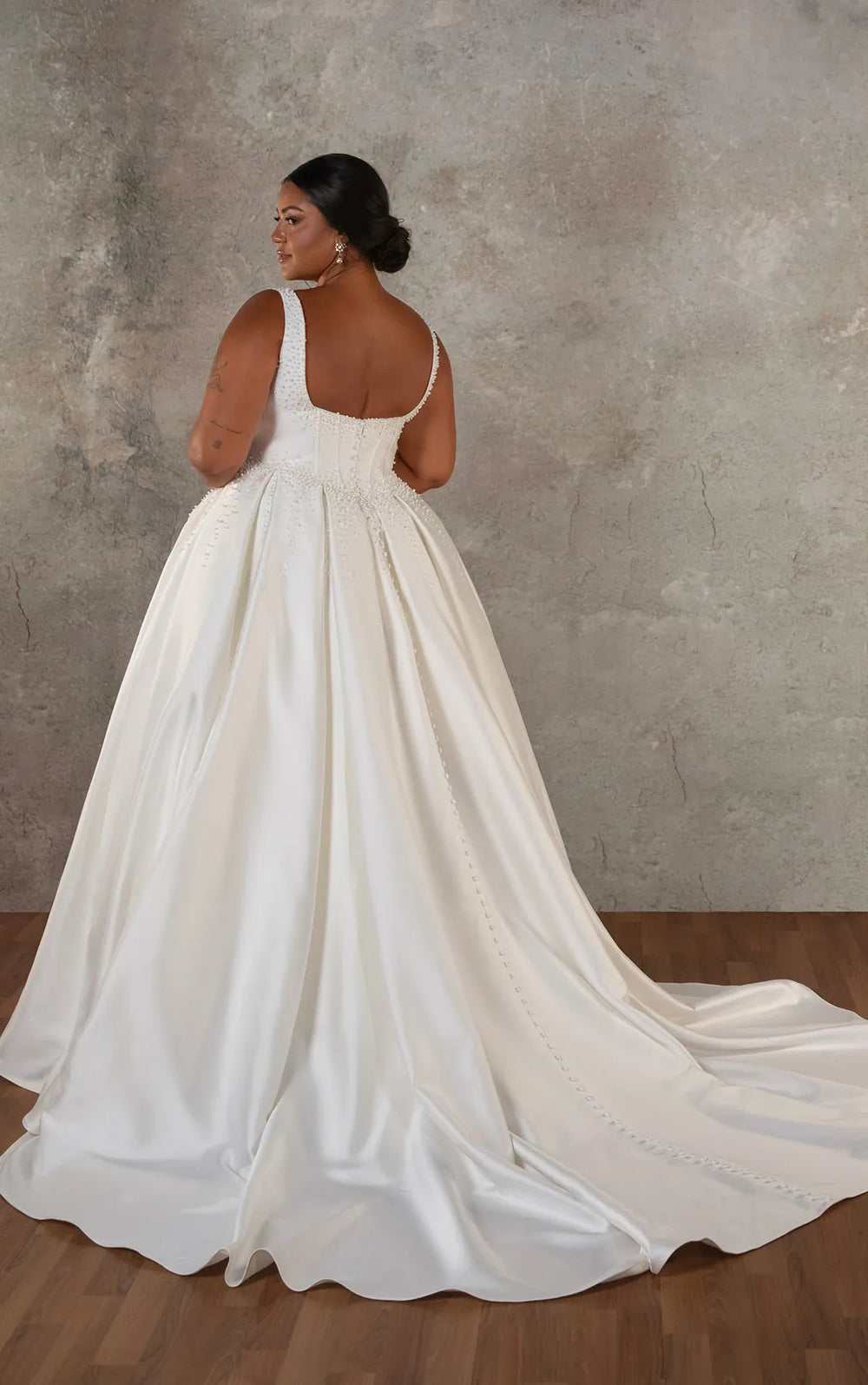 A woman modeling a white satin wedding gown from the back, showing a structured bodice, open back, and full train, set against a textured gray wall and wood floor.