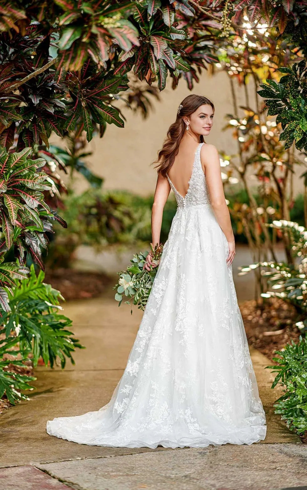 A woman wearing a white lace wedding gown with a deep V back and full train, standing outdoors on a stone path surrounded by lush greenery and warm string lights.
