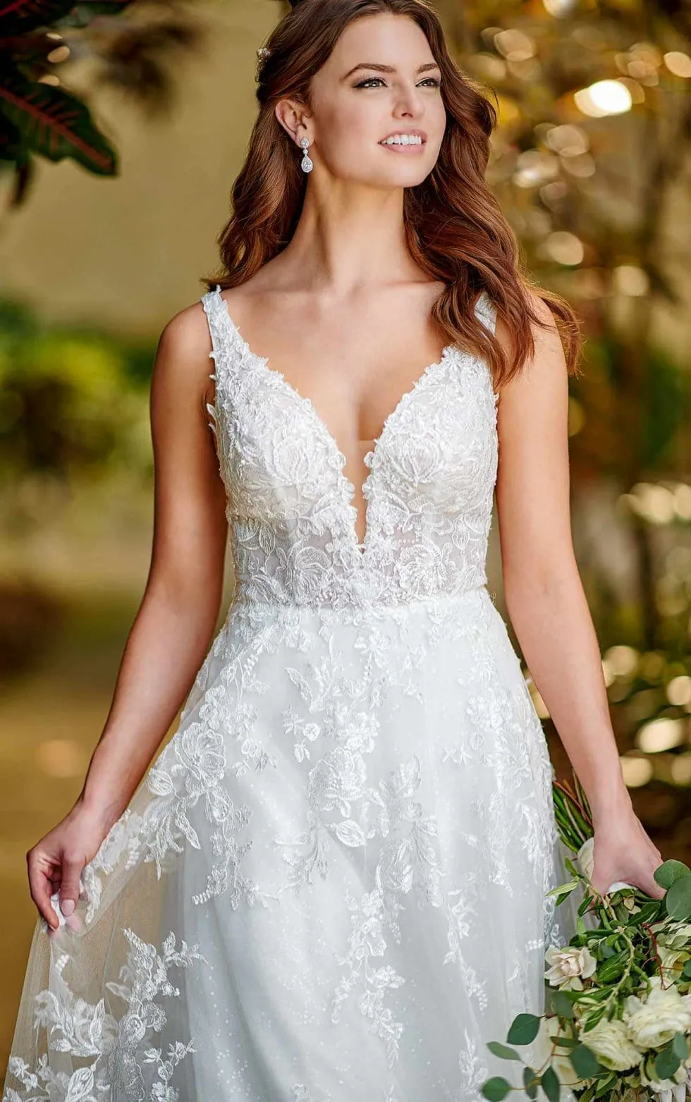 A woman wearing a white lace wedding dress with a deep V-neckline and sheer floral embroidery, holding a bouquet of white flowers in a softly blurred outdoor setting.