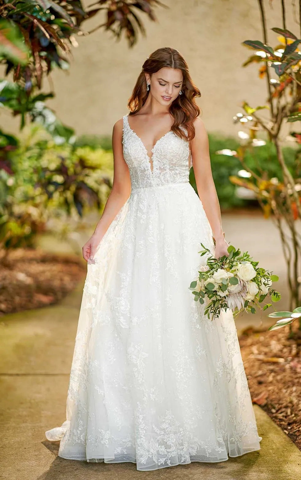 A woman wearing a white lace A-line wedding dress with a deep V-neckline and holding a bouquet of white flowers, standing outdoors on a stone path surrounded by green foliage.