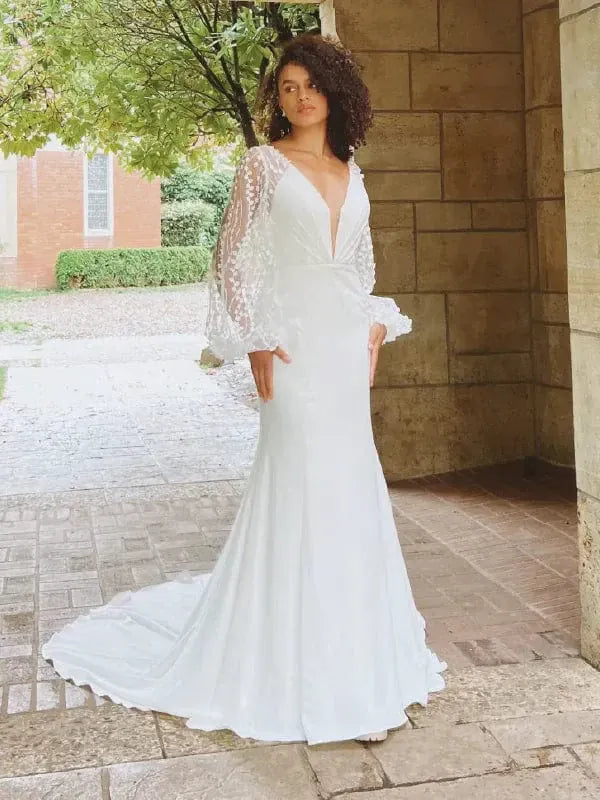 A woman wearing a white mermaid-style wedding gown with a deep V-neckline, sheer lace long sleeves, and a slight train, standing outdoors against a stone wall and brick building background.