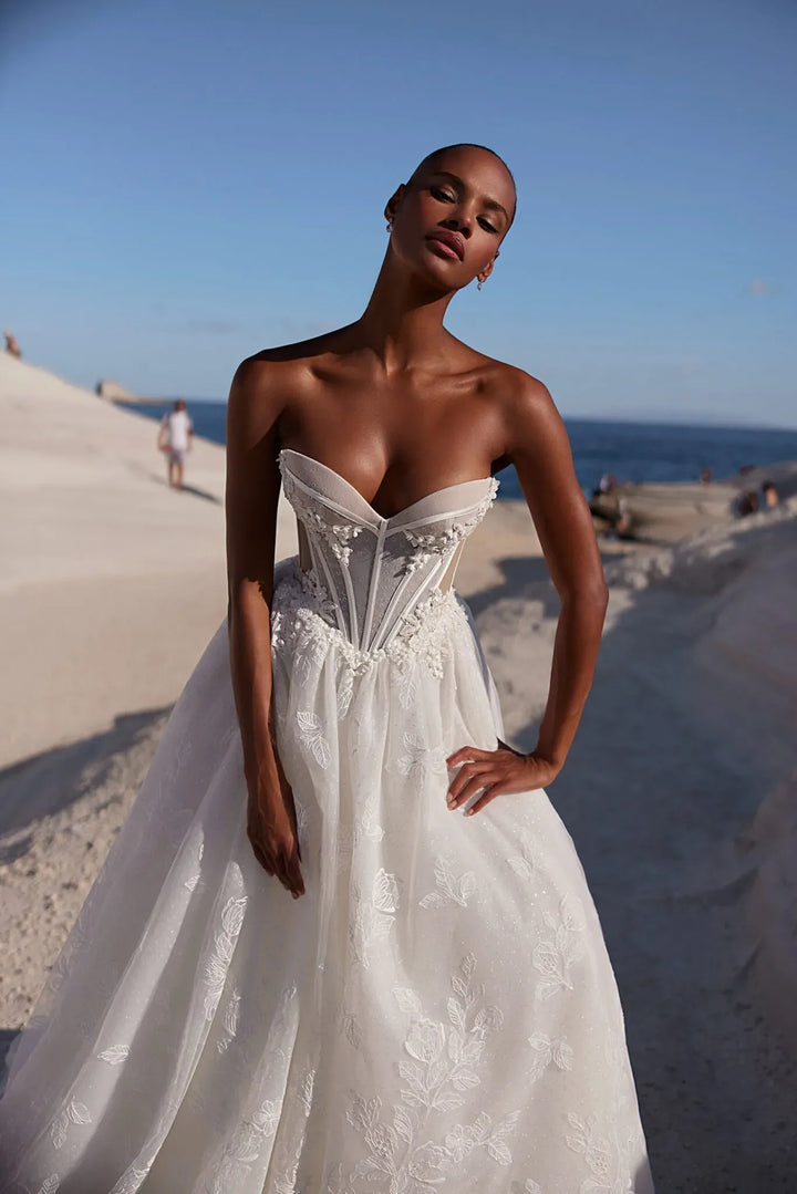 A woman wearing a strapless white wedding gown with a structured corset-style bodice and full tulle skirt, standing on a sandy beach with the ocean and blue sky in the background.