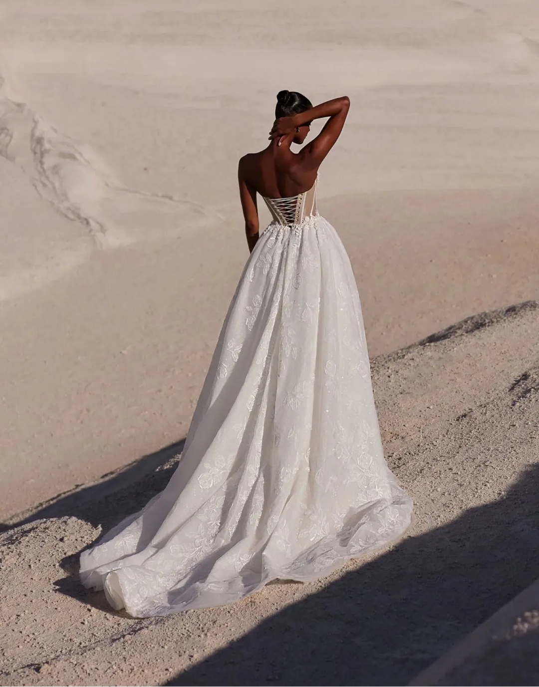 A woman stands on a sandy beach dune wearing a flowing white wedding dress, photographed from behind as she looks out toward the ocean.