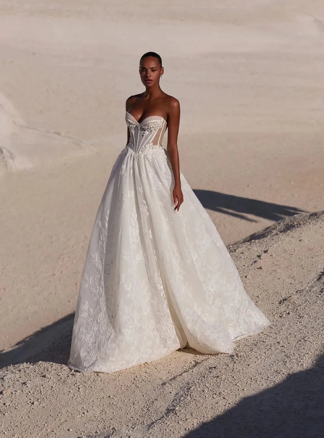 A woman wearing a strapless white wedding gown stands on sandy dunes outdoors, with the ocean and a rock visible in the background.
