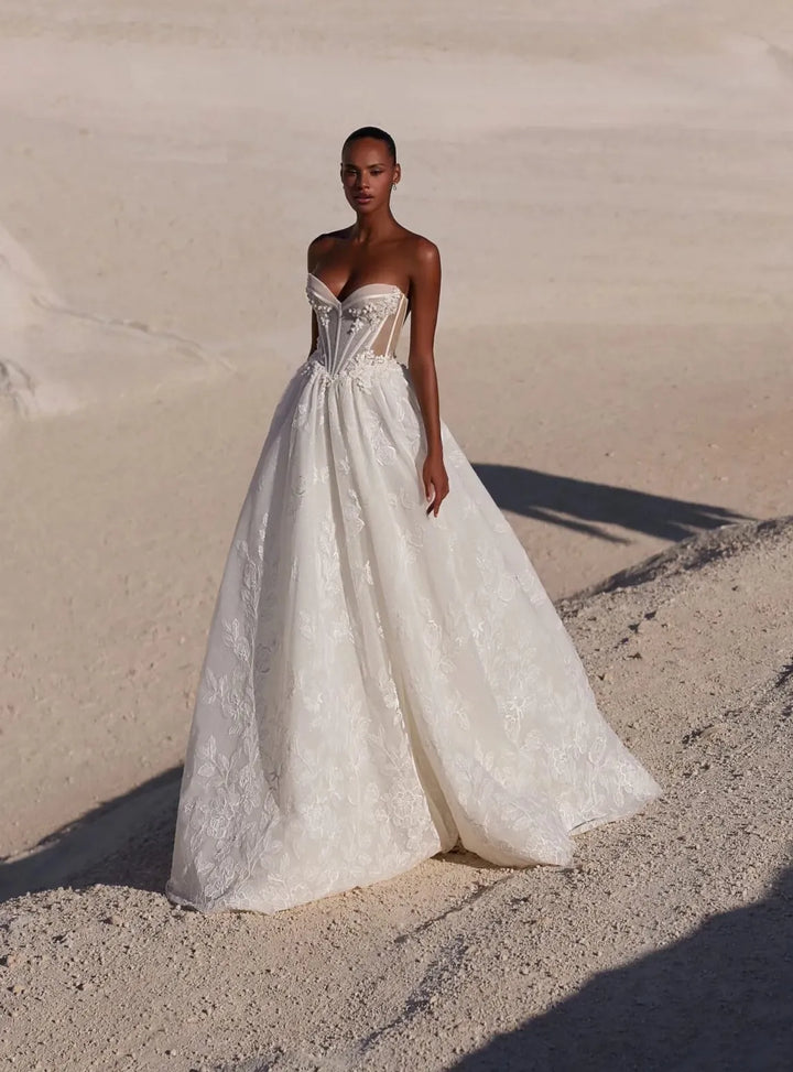 A woman wearing a strapless white wedding gown stands on sandy dunes outdoors, with the ocean and a rock visible in the background.
