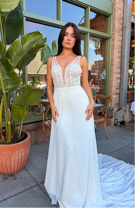 A woman wearing a white sleeveless wedding gown with a lace-embellished bodice and flowing skirt, standing outdoors in front of a storefront with large green-framed windows and a potted plant.