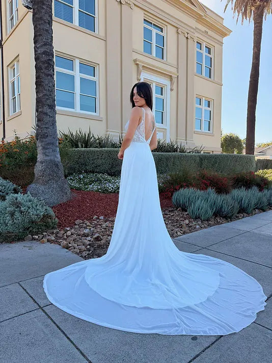 A woman wearing a white wedding dress with a long train stands outdoors on a sidewalk in front of a beige building with palm trees and landscaped plants.