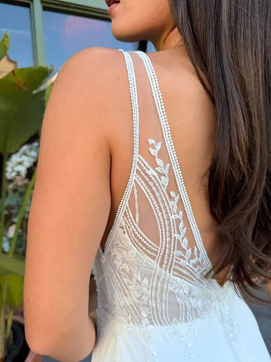Close-up back view of a woman wearing a white lace dress with thin beaded straps and an open back, photographed outdoors near greenery.