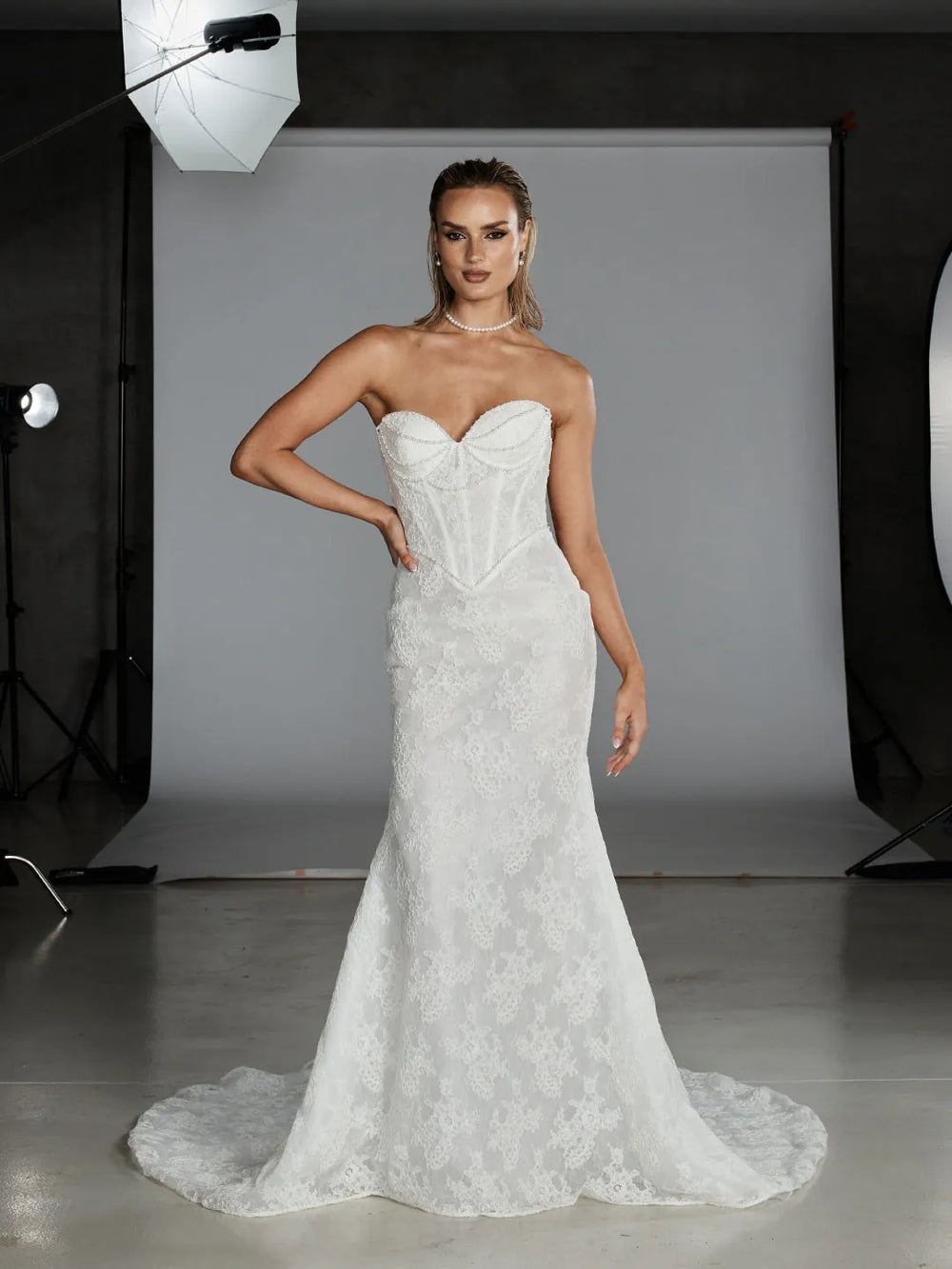 A woman models a strapless white lace mermaid-style wedding gown with a structured corset bodice in a studio setting with visible softbox lights and a gray backdrop.