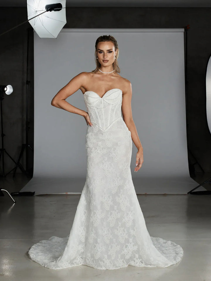 A woman models a strapless white lace mermaid-style wedding gown with a structured corset bodice in a studio setting with visible softbox lights and a gray backdrop.