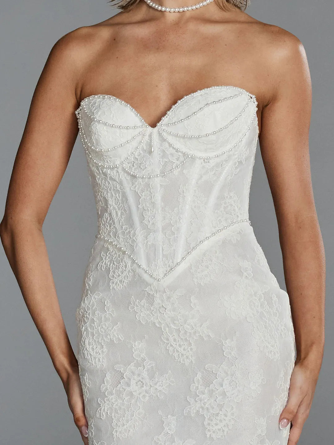 A strapless white lace dress with a sweetheart neckline and beaded corset-style bodice, shown on a model against a gray studio background.
