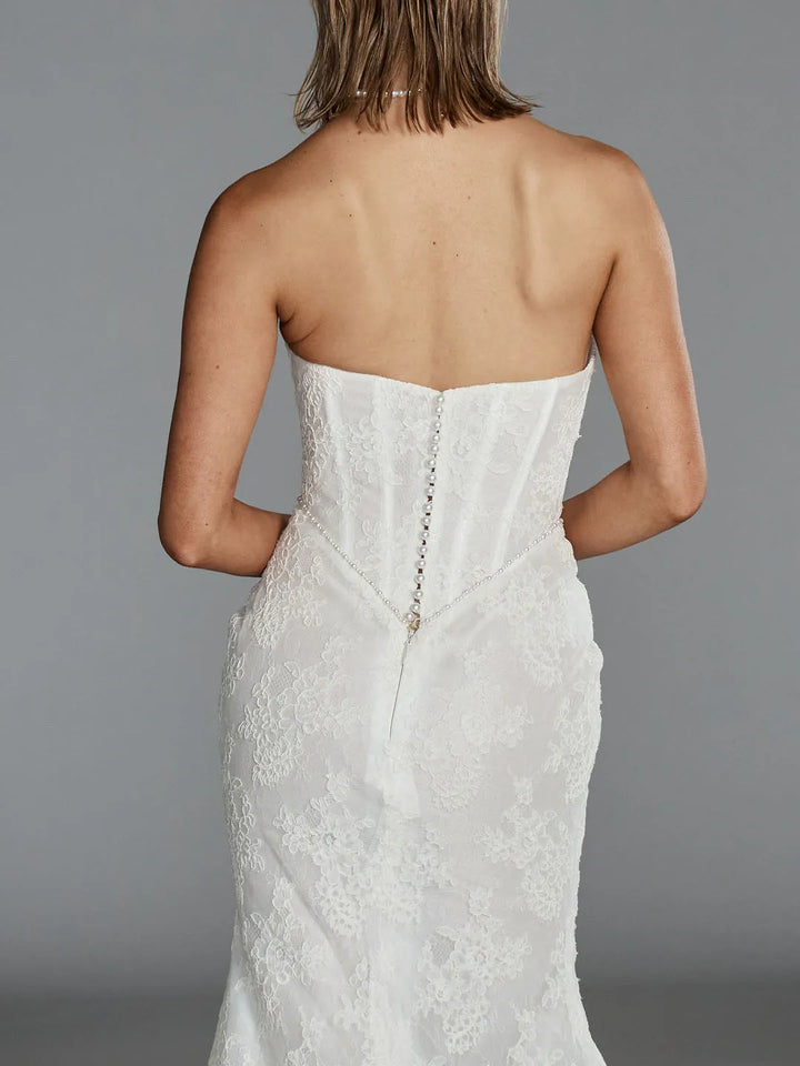 Rear view of a woman wearing a strapless white lace wedding dress with a fitted corset-style back and a central decorative chain detail, against a plain gray studio background.