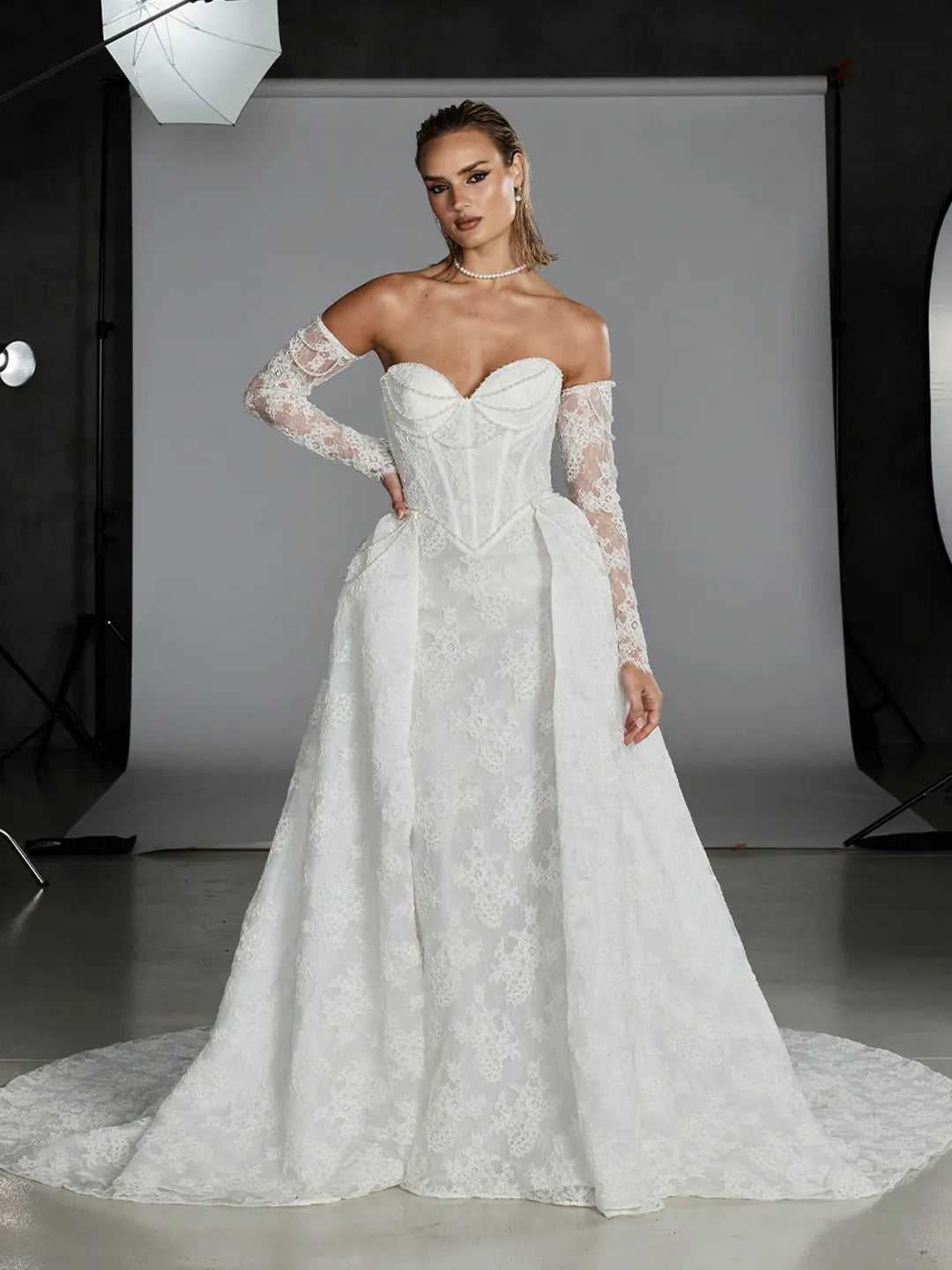 A model wearing a white off-the-shoulder lace wedding gown with a structured corset bodice and long sheer lace sleeves, standing in a studio setting.